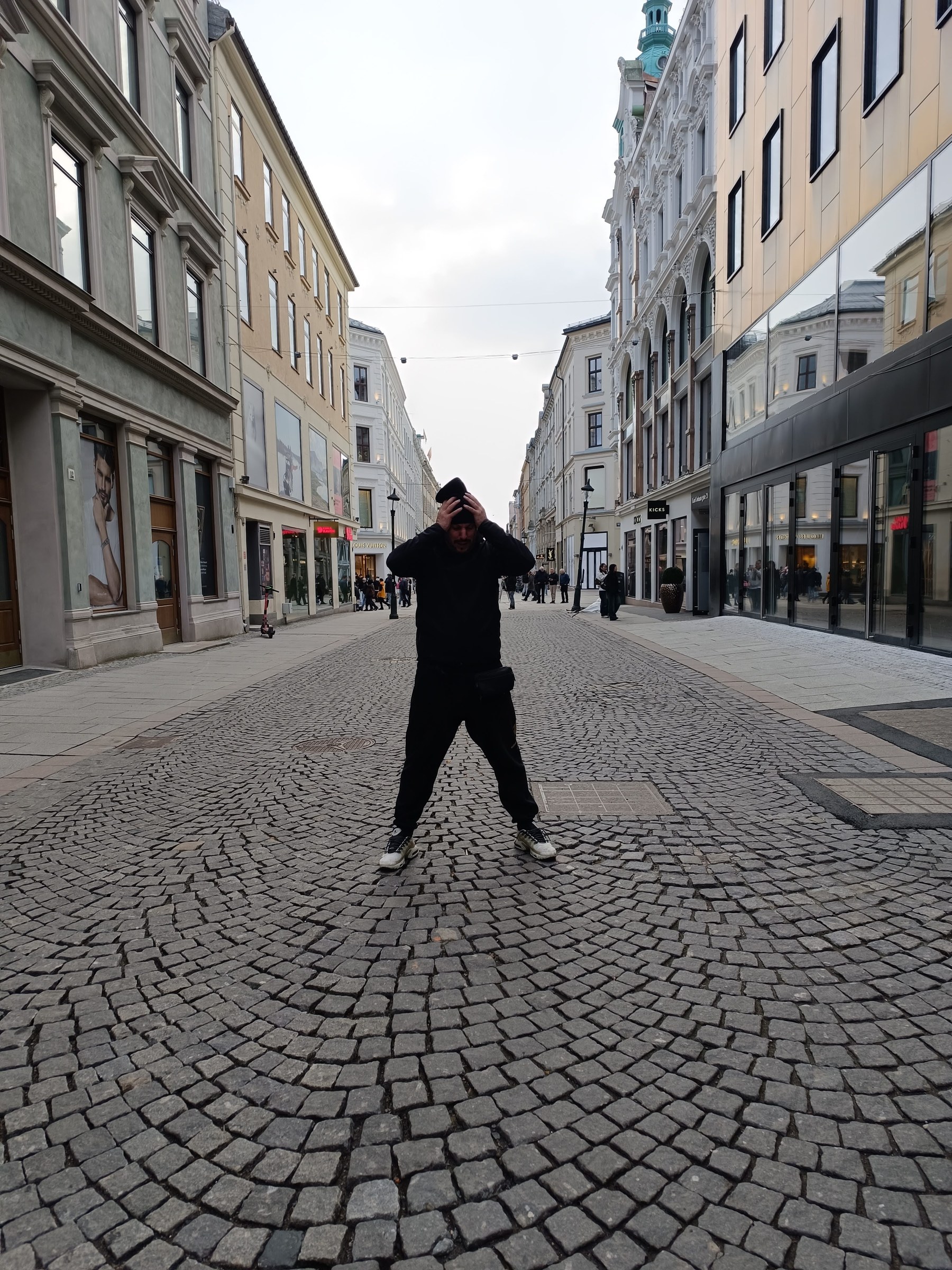 FUNESMA79 de pie en una calle urbana europea, vestido de oscuro, ajustándose el gorro, encuadre de cuerpo entero entre fachadas