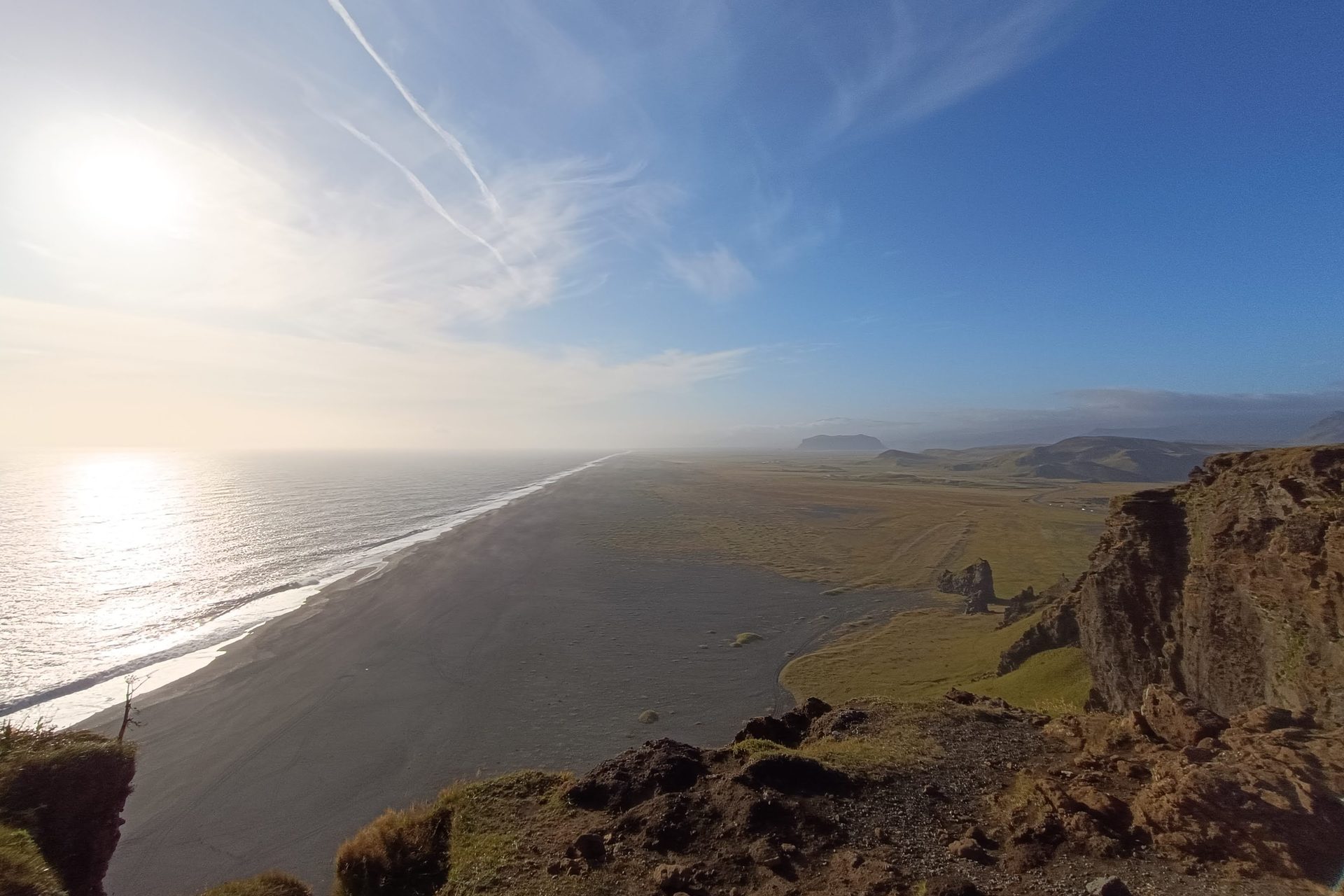 Vista desde un acantilado sobre una playa de arena negra que se extiende hasta el horizonte, con el sol bajo sobre el mar
